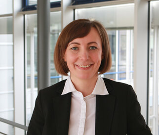 A smiling woman with shoulder-length brown hair is wearing a black blazer and a white shirt. She is standing in a modern office building.
