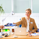 A woman in an office hands a document to a man while sitting at a desk with a laptop and office supplies.