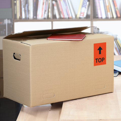 Ein Karton mit dem Aufkleber "FRAGILE", daneben ein graues Kuvert mit "DRINGEND!" und ein weißes Kuvert. Scheren liegen ebenfalls auf dem Tisch.