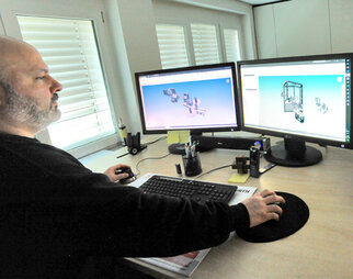 A man is working at a computer with two screens displaying 3D models. The desk is equipped with office supplies.