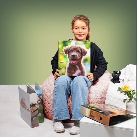 A girl is sitting on a pouf, holding a binder with a picture of a dog. Next to her are more binders and a small plant.