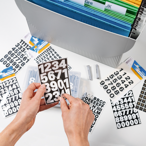 Hands are holding a black board with white numbers. In the background, more numbers and letters, as well as a storage system, are visible.