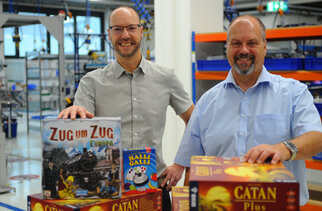 Two men are standing in a toy store, showcasing board games, including 'Ticket to Ride: Europe,' 'Halli Galli,' and 'Catan Plus.