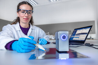 A person in a lab coat and safety goggles is using a marker to write on a shiny surface, while a laptop sits next to them.