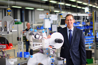 A man in a suit stands next to a machine in a factory, while other employees are busy in the background.