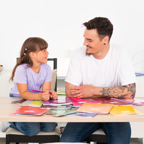 Ein Mann und ein Kind sitzen an einem Tisch, umgeben von bunten Farbmusterkarten. Sie scheinen eine kreative Aktivität zu planen.