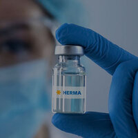 A person wearing blue gloves is holding a small glass bottle labelled 'HERMA'.