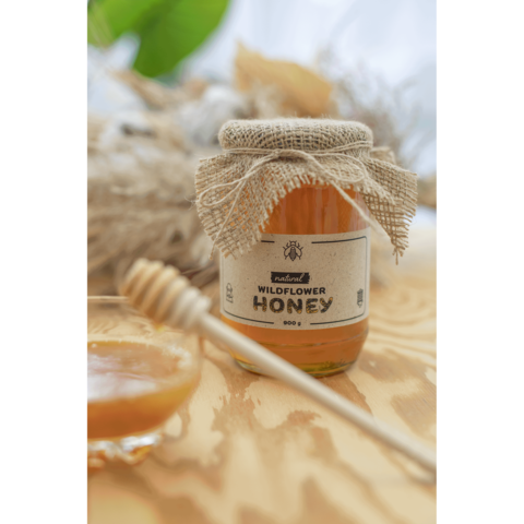 Glas Honig mit Jute-Deckel, beschriftet mit "Wildflower Honey", neben einem kleinen Glas und einem Honiglöffel auf einem Holzuntergrund.