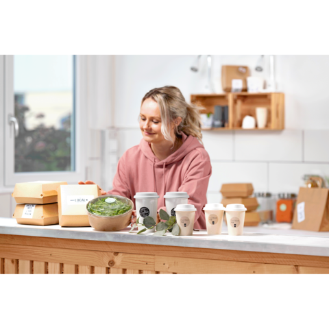 Eine lächelnde Frau in einem rosa Hoodie steht an einem Tisch mit Essensverpackungen, einer Schüssel Salat und Kaffeebechern.