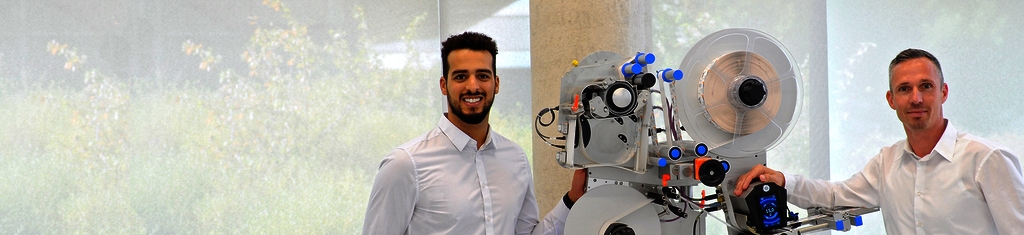 Two men in white shirts are standing next to a technical machine with various components and controls.