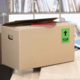 Kartonbox mit einem roten Heft auf dem Deckel, beschriftet mit einem grünen "TOP"-Schild. Im Hintergrund sind Bücherregale sichtbar.