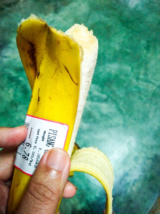 A hand is holding a partially peeled banana with a price tag indicating the weight and price.