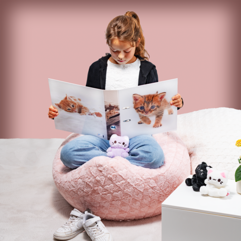 A girl is sitting in a large, soft bean bag and looking at a book filled with pictures of kittens. Stuffed animals are scattered around her.