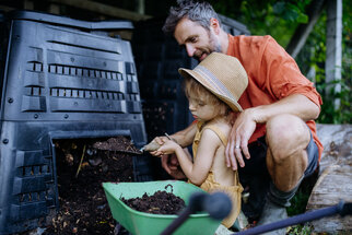 An adult and a child are shoveling compost from a container into a wheelbarrow. Both are wearing practical clothing and a sun hat.