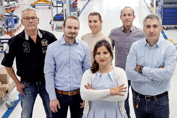 A group of seven people stands in a factory hall, surrounded by machines and workstations.