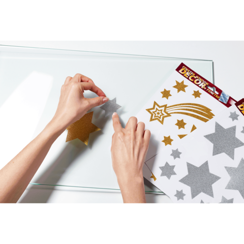 Eine Hand platziert einen glitzernden Stern auf einer transparenten Oberfläche, während ein Bogen mit weiteren glitzernden Sternen und Motiven daneben liegt.
