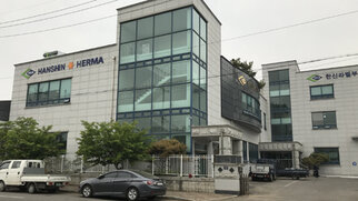 Modern office buildings with large windows, labelled 'HANSHIN' and 'HERMA'. Surrounded by trees and parked cars.