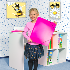 Ein Mädchen mit blonden Locken hält ein großes pinkes Blatt Papier in einem bunten Raum mit Bienenmotiven an den Wänden.