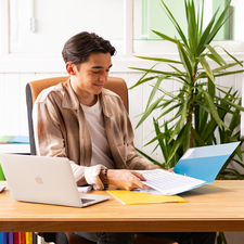 Ein junger Mann sitzt an einem Schreibtisch, arbeitet mit einem Laptop und blättert durch Dokumente in einer Ordnermappe. Pflanzen und farbige Akten sind im Hintergrund.
