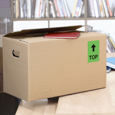 Kartonbox auf einem Tisch, mit einem roten Heftchen oben und einem grünen "TOP"-Schild. Hintergrund zeigt Bücherregale.