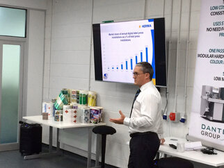 A man is presenting in front of a screen displaying a chart about digital printing machines. Various printing materials are visible in the background.