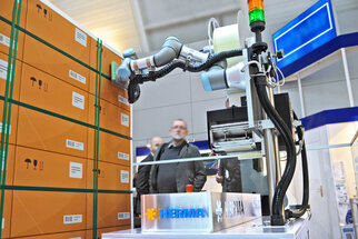 Industrial robot from Herrmann interacting with boxes in a showroom, with a sign displaying the company name in the foreground.