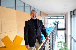 A man with glasses and gray hair stands at a railing in a modern building featuring wooden and plant elements.
