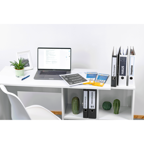 Ein Schreibtisch mit einem Laptop, mehreren Ordnern, Dokumenten und Pflanzen. Im Hintergrund ein Bilderrahmen.