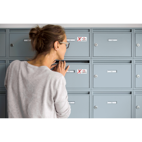 Eine Frau schaut in einen grauen Briefkasten mit einem roten X-Symbol. Die Umgebung zeigt mehrere ähnliche Briefkästen.