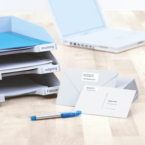 Ordner mit beschrifteten Fächern für eingehende und ausgehende Dokumente, dazu Briefumschläge und ein blauer Stift auf einem Tisch.