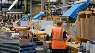 Workers in an orange vest sorting boxes and interacting with automated systems in a warehouse.