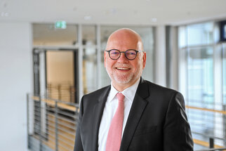 A smiling man in glasses and a suit stands in a modern interior. He is wearing a pink tie and has a friendly demeanor.