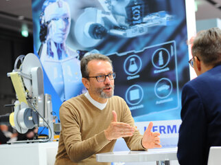 A man in conversation with another, while a technical presentation and a robot are visible in the background.