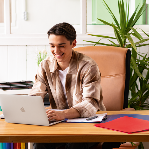 Ein junger Mann sitzt an einem Schreibtisch mit einem Laptop, umgeben von Büromaterialien und Pflanzen, und arbeitet konzentriert.