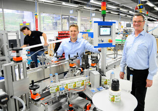Two men are standing at a production line in a factory, while a third worker is working in the background.