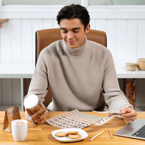 Ein junger Mann sitzt an einem Tisch, betrachtet ein Blatt mit Mustern und hält ein Glas mit Keksen in der Hand. Vor ihm stehen eine Tasse Kaffee und ein Laptop.