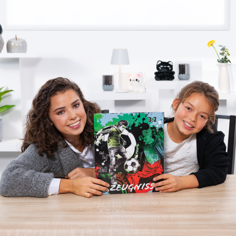 Two girls are holding a soccer report card with a graphic and a score of 3:2. They are sitting at a table in a bright room.
