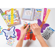 Bunte Aufkleber, Schmetterlinge und ein Notizbuch liegen auf einem Tisch. Hände fügen Sticker hinzu. Bücher und weitere Sticker sind im Hintergrund sichtbar.