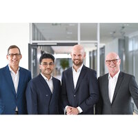 Four men in suits are standing in a modern office hallway. They are smiling and posing next to each other.