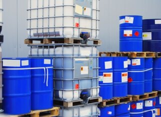 Stacked blue barrels and containers on pallets, marked with various labels and warning signs.