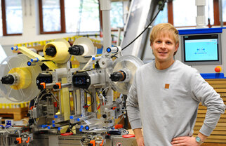 A man stands next to a modern machinery setup for processing materials, surrounded by technical devices and screens.