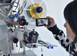 A person is holding a smartphone and taking a picture of a machine with various technical components and wheels.