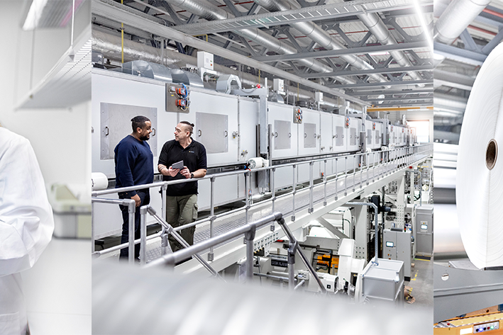 Two women in lab coats are examining a document, while in the background, a production line with large rolls can be seen.