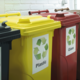 Recyclingbehälter in verschiedenen Farben: Gelb für Plastik, Rot für Glas und Schwarz für andere Materialien, mit entsprechenden Symbolen.