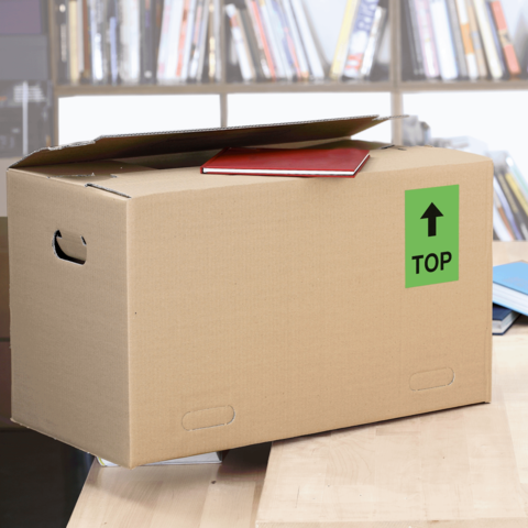 Kartonbox auf einem Tisch, mit einem roten Heft und einem grünen "TOP"-Schild auf der Seite. Hintergrund mit Bücherregalen.