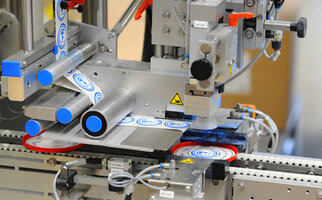 A machine component that applies labels to round objects, featuring various mechanical parts and a conveyor belt in the background.