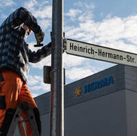 A worker is attaching a street sign reading 'Heinrich-Hermann Street' to a post, while a building is visible in the background.