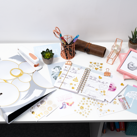 Bürotisch mit Schreibwaren, Notizbuch, Aufklebern und einer kleinen Pflanze. Verschiedene Bastelmaterialien sind verteilt.