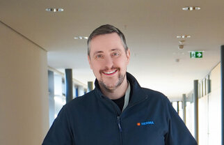 A man with short hair and a beard is wearing a black jacket with the HERMA logo. He is standing in a modern hallway.