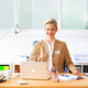 A woman with blonde hair is sitting at a desk, smiling and wearing a blue blazer. In front of her is a laptop and some writing materials.